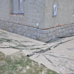 Fragment elewacji budynku z nową kamienną podmurówką. Okna zabezpieczone folią, teren wokół budynku osłonięty plandeką. Widoczne ślady prac remontowych.