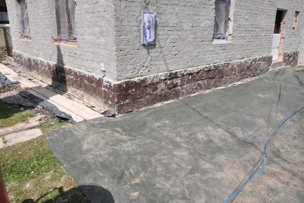 Fragment elewacji budynku z zabezpieczonymi oknami i plandeką ochronną, ukazujący etap prac renowacyjnych dolnej części ściany.