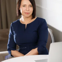 Portret kobiety w granatowej sukience siedzącej przy białym stole z laptopem, patrzącej w obiektyw.