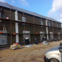 Rd-Complex Rafal Dettlaff - Dwóch pracowników na rusztowaniu podczas nakładania tynku na elewację budynku z oknami zabezpieczonymi folią, widoczne materiały budowlane i worki na śmieci.