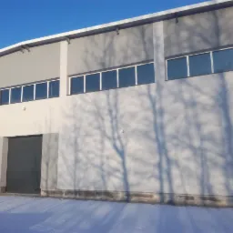 Świeżo otynkowana elewacja hali przemysłowej w zimowej scenerii, z widocznymi cieniami drzew na fasadzie i częściowo ośnieżonym terenem.