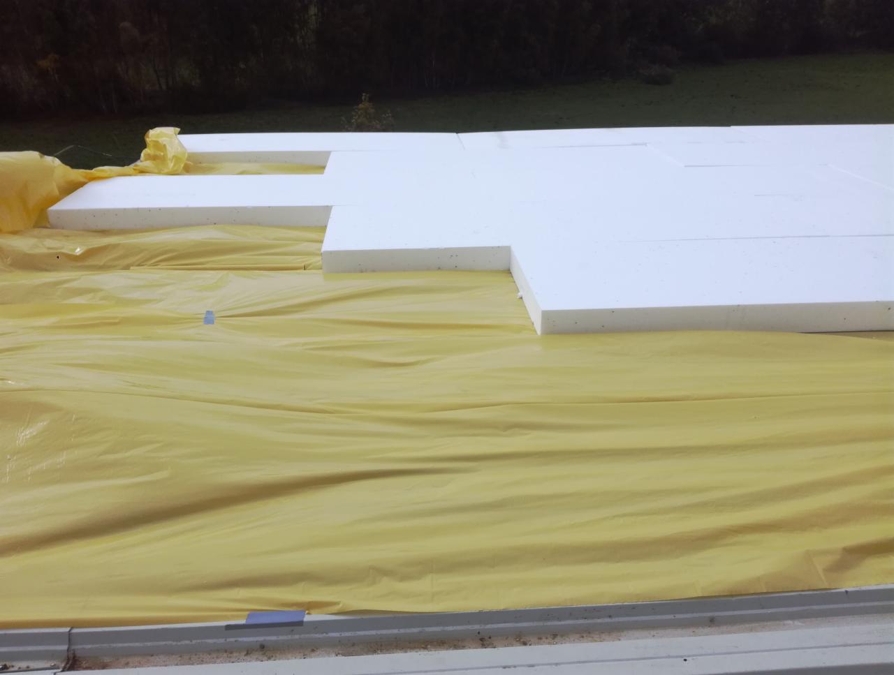 Układanie białych płyt styropianowych na żółtej folii izolacyjnej, widok z góry na częściowo ocieplony dach.