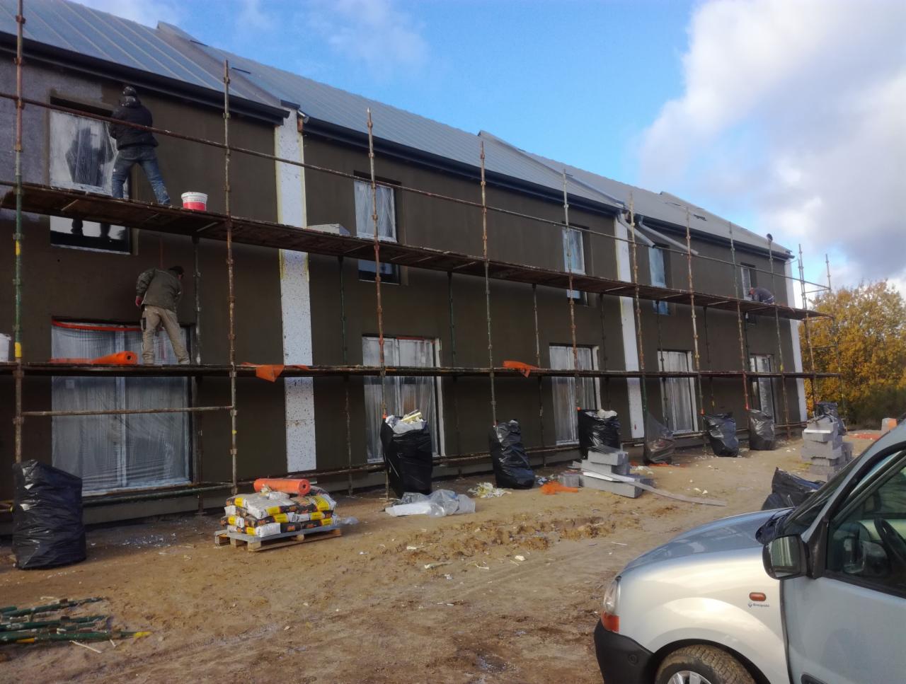 Dwóch pracowników na rusztowaniu podczas nakładania tynku na elewację budynku z oknami zabezpieczonymi folią, widoczne materiały budowlane i worki na śmieci.