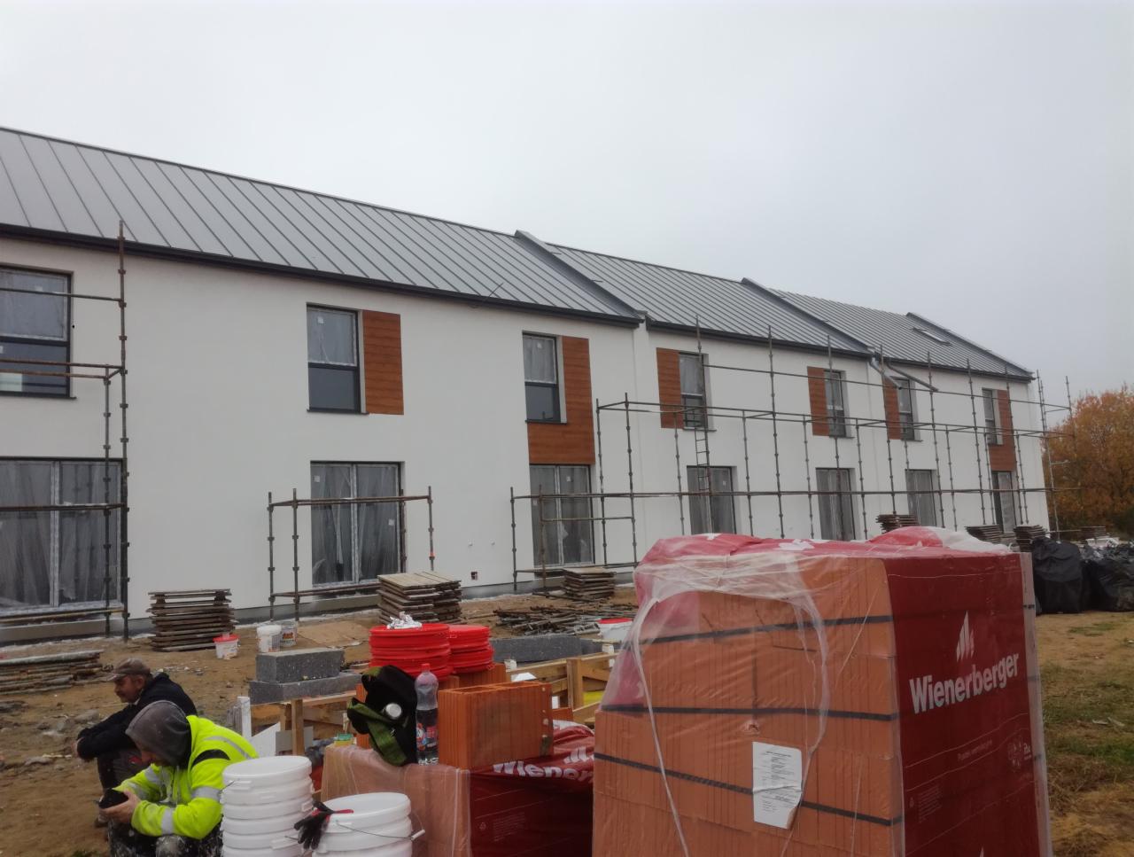 Budynek w trakcie prac elewacyjnych: biała fasada, drewniane panele dekoracyjne, rusztowania, palety z cegłami Wienerberger, pracownicy w odblaskowych kamizelkach.