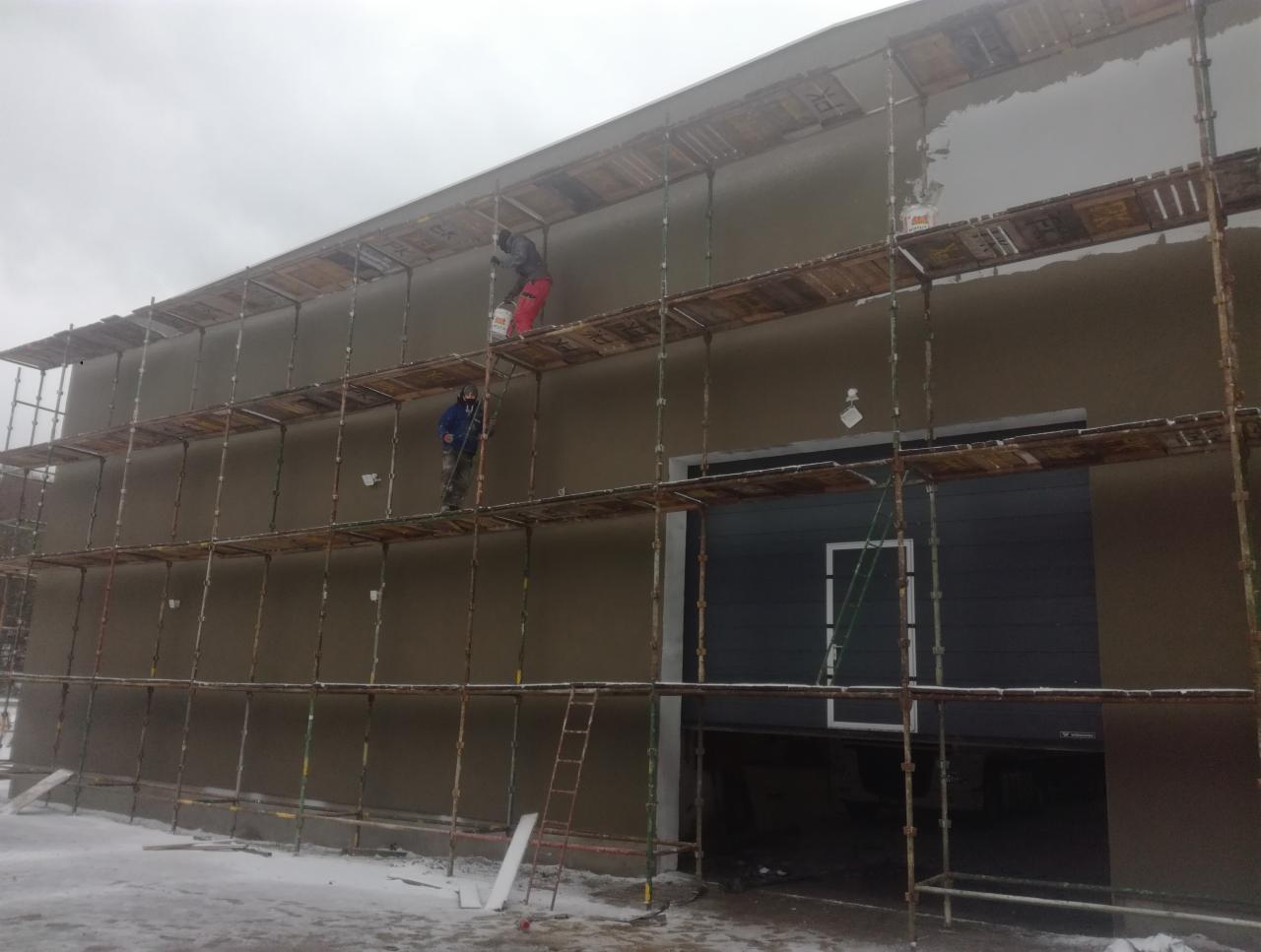 Dwóch pracowników na rusztowaniu podczas nakładania tynku na ścianę budynku w pochmurny dzień zimowy, widoczne ślady śniegu na ziemi.