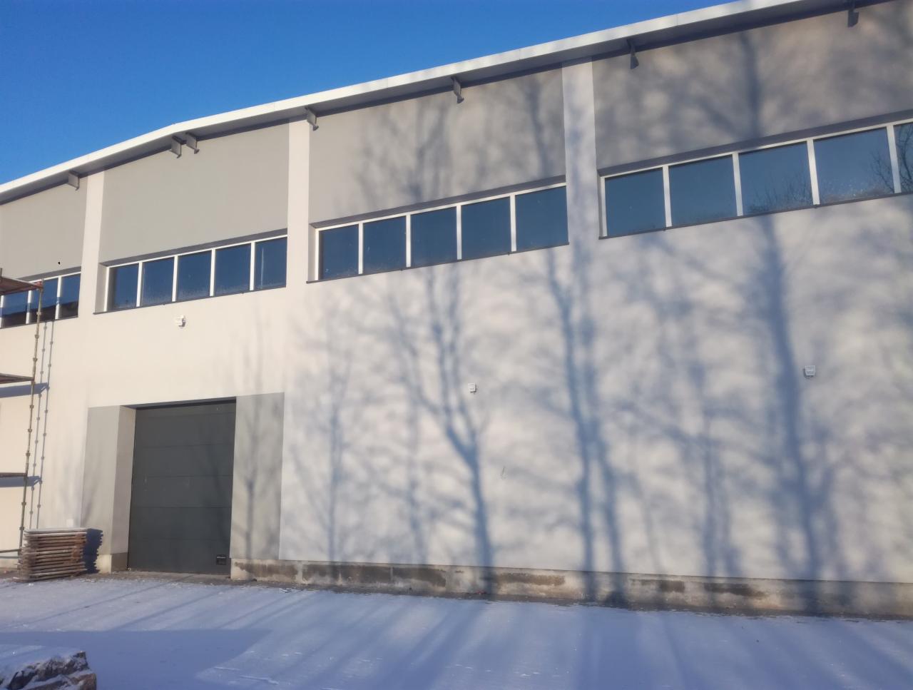 Świeżo otynkowana elewacja hali przemysłowej w zimowej scenerii, z widocznymi cieniami drzew na fasadzie i częściowo ośnieżonym terenem.