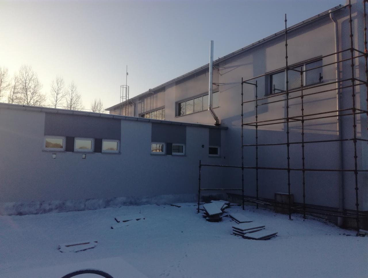 Budynek z szarą elewacją w zimowej scenerii, widoczne rusztowanie i komin na ścianie, śnieg zalega na ziemi.