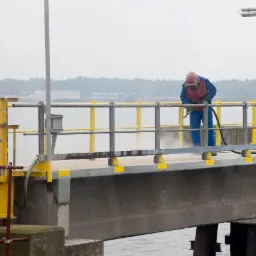 Pracownik w kombinezonie ochronnym czyści metalową konstrukcję mostu za pomocą strumienia ścierniwa. Konstrukcja pomalowana na żółto. W tle woda i zalesiony brzeg.