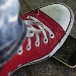 Czerwony tenisówka depcze smartfon z pękniętym ekranem, widoczne jeansy, szare tło.