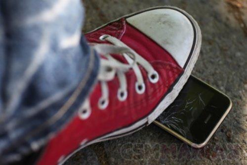 Czerwony tenisówka depcze smartfon z pękniętym ekranem, widoczne jeansy, szare tło.