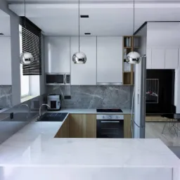 Nowoczesna kuchnia z marmurowym blatem, białymi szafkami i drewnianymi akcentami, oświetlona trzema kulistymi lampami wiszącymi, widok na salon z czarnym telewizorem.