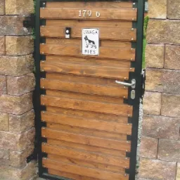 Drewniana furtka z numerem domu i tabliczką ostrzegawczą 'Uwaga pies', osadzona w kamiennym ogrodzeniu, z widoczną kostką brukową u podstawy.