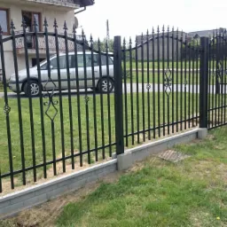 Czarne ogrodzenie kute z dekoracyjnymi elementami i podmurówką z szarej cegły, chroniące posesję z widocznym samochodem w tle.