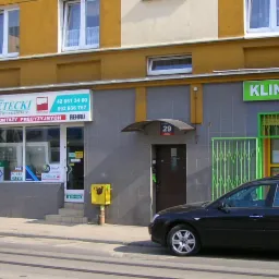 Fasada budynku z oknami i drzwiami PCV/Aluminium, szyldem 'Firma Petecki'. Widoczny fragment ulicy z czarnym samochodem osobowym. Dzień, światło naturalne.