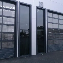 Zewnętrzny widok trzech okien o różnych rozmiarach i konstrukcjach: dwa segmentowe z wieloma szybami i jedno wąskie, wysokie, bez podziałów, wszystkie w ciemnych ramach, wbudowane w szarą ścianę...