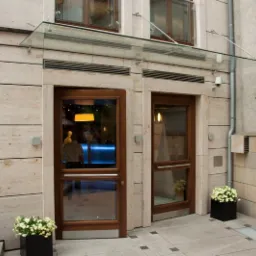 Dwa wejścia do budynku z drewnianymi drzwiami z przeszkleniami, nad drzwiami szklany daszek, elewacja z kamienia, czarne donice z białymi kwiatami po bokach, w tle widoczne okna z kwiatami.