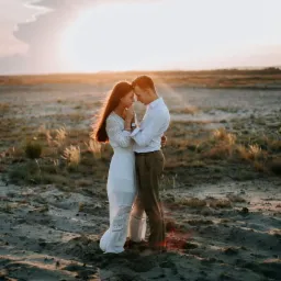 Para narzeczonych w objęciach na piaszczystym terenie o zachodzie słońca, kobieta w białej sukience z koronkowymi wstawkami, mężczyzna w białej koszuli i brązowych spodniach.