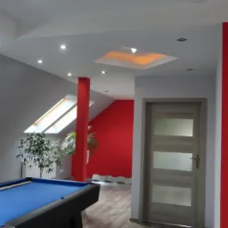 Pokój na poddaszu z oknami dachowymi, pomalowany na szaro i czerwono. Na pierwszym planie stół bilardowy z niebieskim suknem. Widoczne drzwi wewnętrzne.