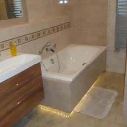 Jasna łazienka z brązową szafką pod umywalką, wanną z podświetleniem LED i beżowymi płytkami. Widoczny grzejnik drabinkowy i lustro.