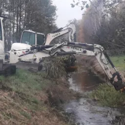 Biała koparka Terex TC75 z operatorem, usuwająca roślinność z rowu melioracyjnego w pochmurny dzień.
