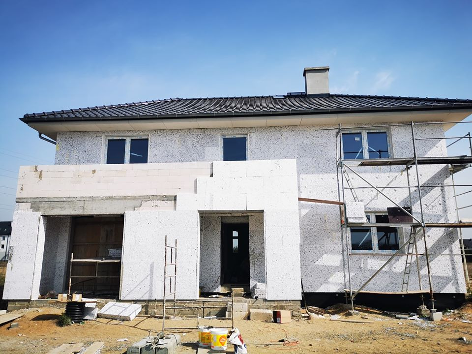 Dom jednorodzinny w trakcie prac elewacyjnych, z widocznymi rusztowaniami, oknami i fragmentami ocieplenia, na tle błękitnego nieba.