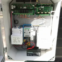 Wnętrze szarej skrzynki z elementami elektronicznymi, w tym płytkami drukowanymi, transformatorem, bezpiecznikami i akumulatorem, zamontowanymi na perforowanej płycie montażowej.