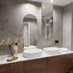 Nowoczesna łazienka z dwoma owalnymi umywalkami nablatowymi, złotymi bateriami i drewnianą szafką pod umywalką, uzupełniona dekoracjami w postaci wazonu z suchymi kwiatami i świeczki na złotej tacy.