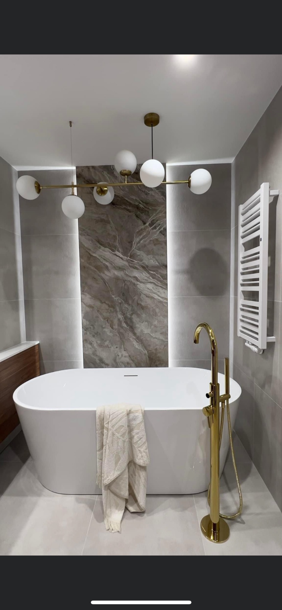 Nowoczesna łazienka z wolnostojącą białą wanną, złotą armaturą i designerską lampą sufitową z białymi kulami, wykończona szarymi płytkami imitującymi kamień.