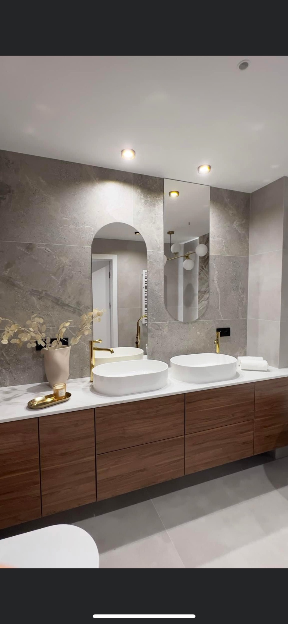 Nowoczesna łazienka z dwoma owalnymi umywalkami nablatowymi, złotymi bateriami i drewnianą szafką pod umywalką, uzupełniona dekoracjami w postaci wazonu z suchymi kwiatami i świeczki na złotej tacy.