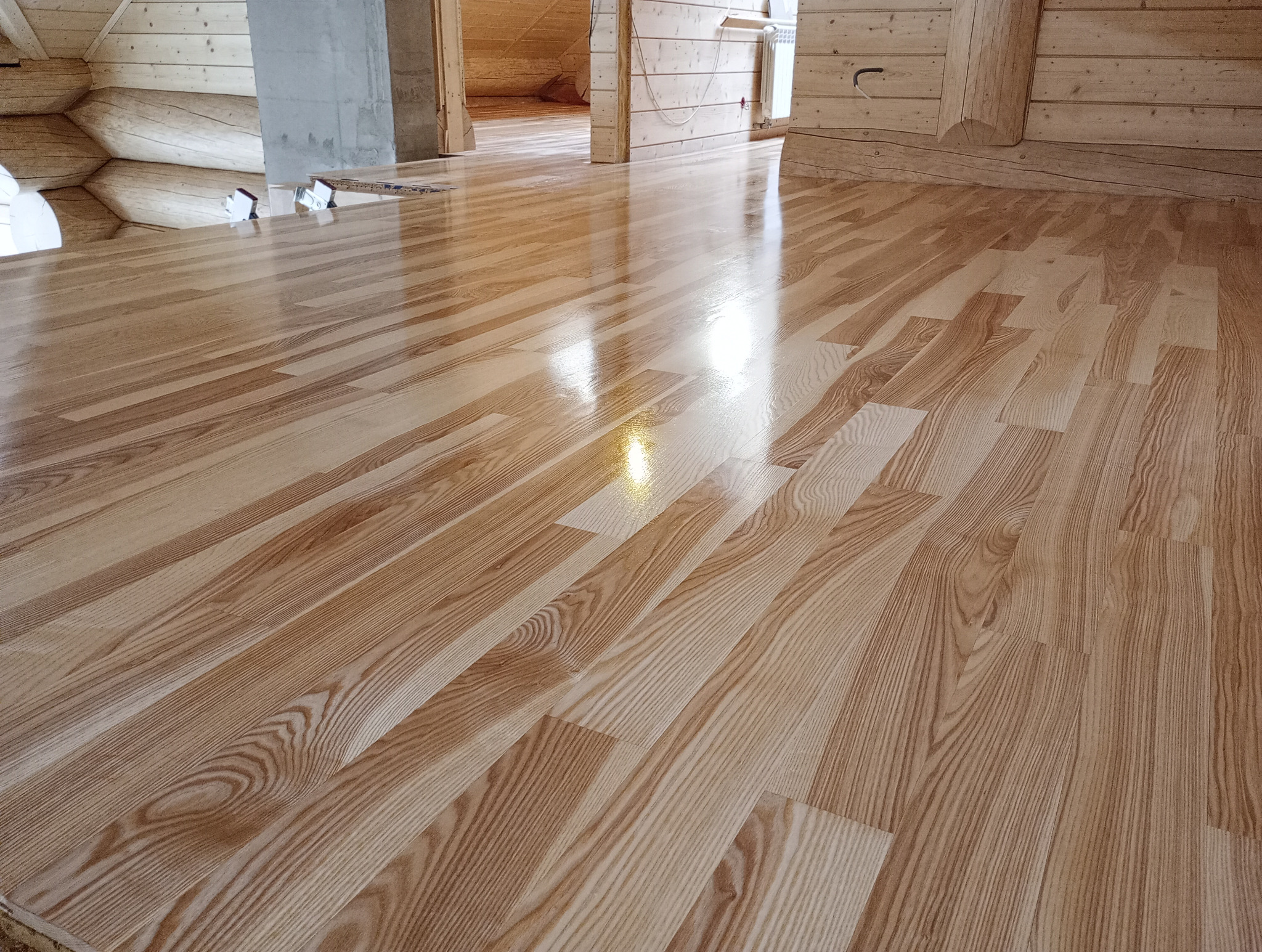 Błyszcząca, jasna podłoga z desek w nowym domu z bali. Widoczne detale drewna i odbicia światła. W tle surowy betonowy filar i drewniane ściany.