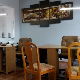 Biuro z drewnianymi meblami: biurka, krzesła z wzorzystymi siedziskami, na ścianie obraz z motywami roślinnymi i kalendarz.