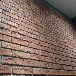 Dekor House Michał Janeczek - Ceglana ściana w pomieszczeniu, wykonana z czerwonej cegły klinkierowej. Widoczny fragment sufitu i narożnik ściany. Układ cegieł w tradycyjnej wiazaniu.
