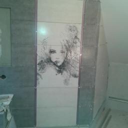 Dekor House Michał Janeczek - Ściana w trakcie wykańczania z szarych i kremowych płytek. Centralnie umieszczony wzór kobiety. Widoczne elementy montażowe i narzędzia.