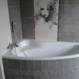 Wykończona łazienka: narożna wanna obudowana szarymi płytkami z mozaiką, chromowana bateria i prysznic, na ścianie płytki z grafiką kobiety. Estetyczne detale.