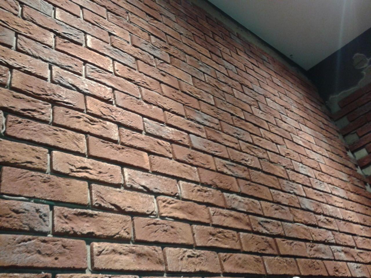 Ceglana ściana w pomieszczeniu, wykonana z czerwonej cegły klinkierowej. Widoczny fragment sufitu i narożnik ściany. Układ cegieł w tradycyjnej wiazaniu.