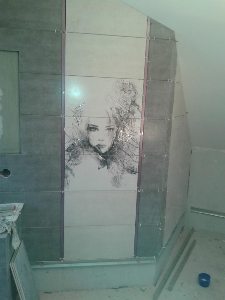 Ściana w trakcie wykańczania z szarych i kremowych płytek. Centralnie umieszczony wzór kobiety. Widoczne elementy montażowe i narzędzia.