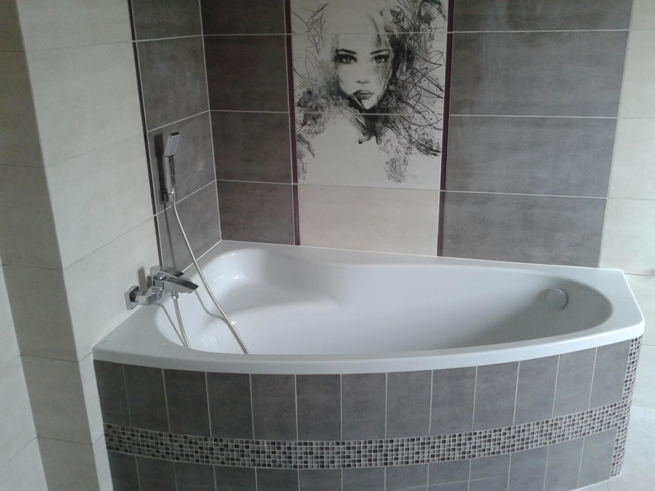 Wykończona łazienka: narożna wanna obudowana szarymi płytkami z mozaiką, chromowana bateria i prysznic, na ścianie płytki z grafiką kobiety. Estetyczne detale.