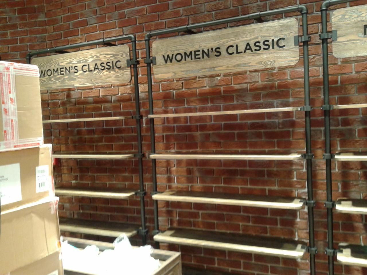 Regał sklepowy z drewnianymi półkami i metalową konstrukcją na tle ściany z czerwonej cegły. Widoczne napisy 'WOMEN'S CLASSIC'. Częściowo zasłonięty stosem kartonów.