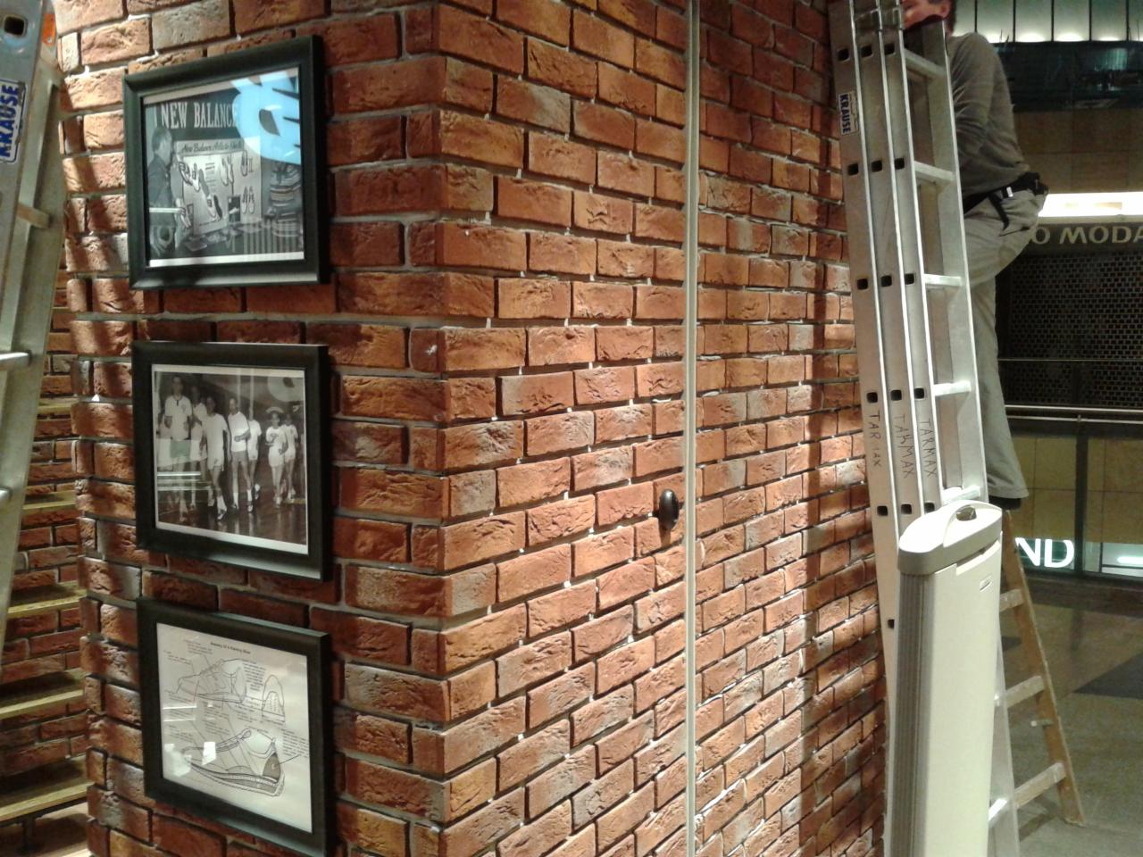 Ściana wyłożona czerwoną cegłą z trzema czarnymi ramkami z grafikami. Obok drabina aluminiowa i fragment sylwetki osoby w szarym ubraniu. Nietypowe wykończenie ściany.