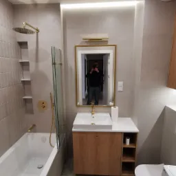 Nowoczesna łazienka z beżowymi płytkami, złotą armaturą, kabiną prysznicową typu walk-in i szafką pod umywalką z drewnianym frontem, widok na odbicie fotografa w lustrze.