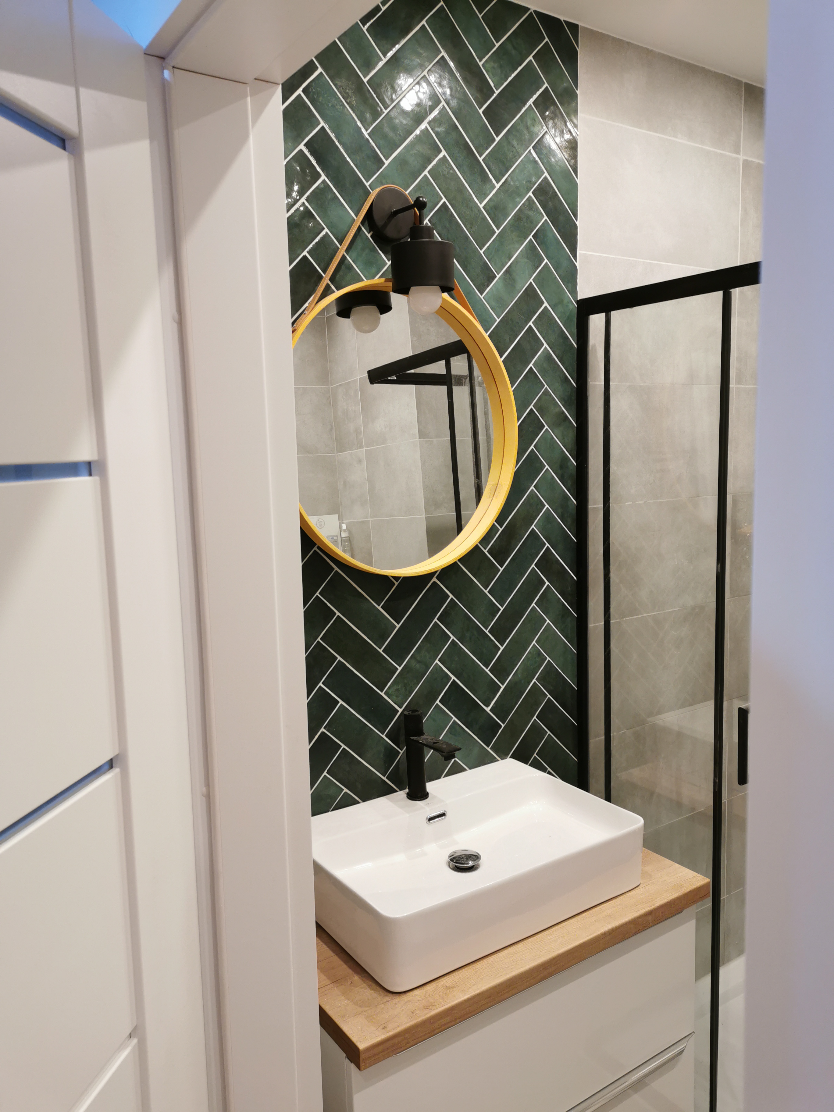 Nowoczesna łazienka z umywalką nablatową, czarną armaturą i lustrem w żółtej ramie na tle zielonych płytek ułożonych w jodełkę.
