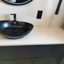 Czarna, owalna umywalka nablatowa z czarną armaturą na jasnym blacie z konglomeratu, widok z góry, minimalistyczny design.