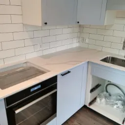 Jasny blat kuchenny z szarymi szafkami, widoczny zlew, płyta indukcyjna w trakcie montażu i otwarte szafki z elementami instalacyjnymi.