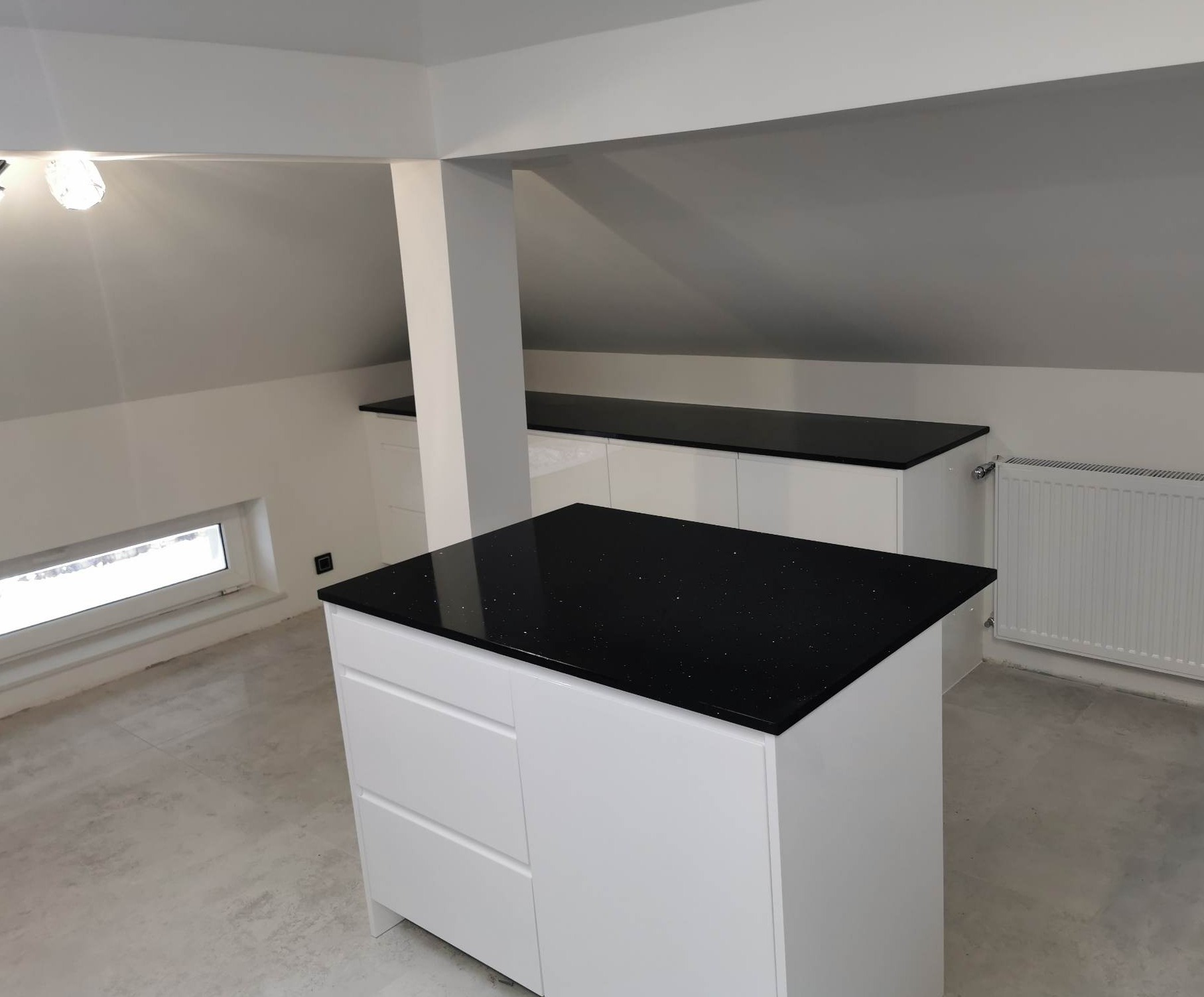 Wyspa kuchenna z czarnym blatem z połyskującymi drobinkami, białe szafki w jasnym pomieszczeniu na poddaszu z oknem i grzejnikiem.