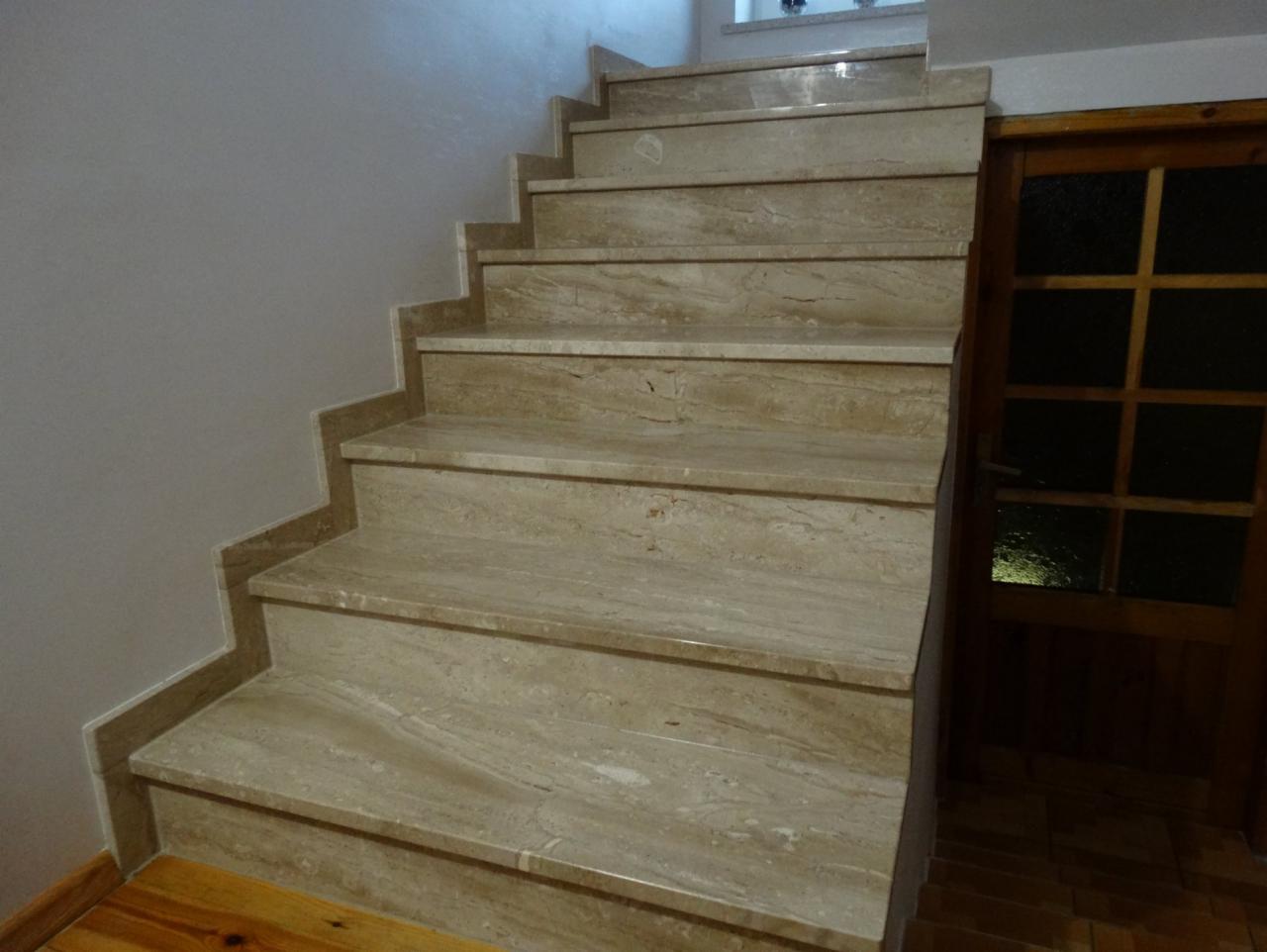 Beżowe schody wewnętrzne z kamienia naturalnego, z widoczną strukturą i usłojeniem, prowadzące do ciemnego pomieszczenia z drewnianymi drzwiami.