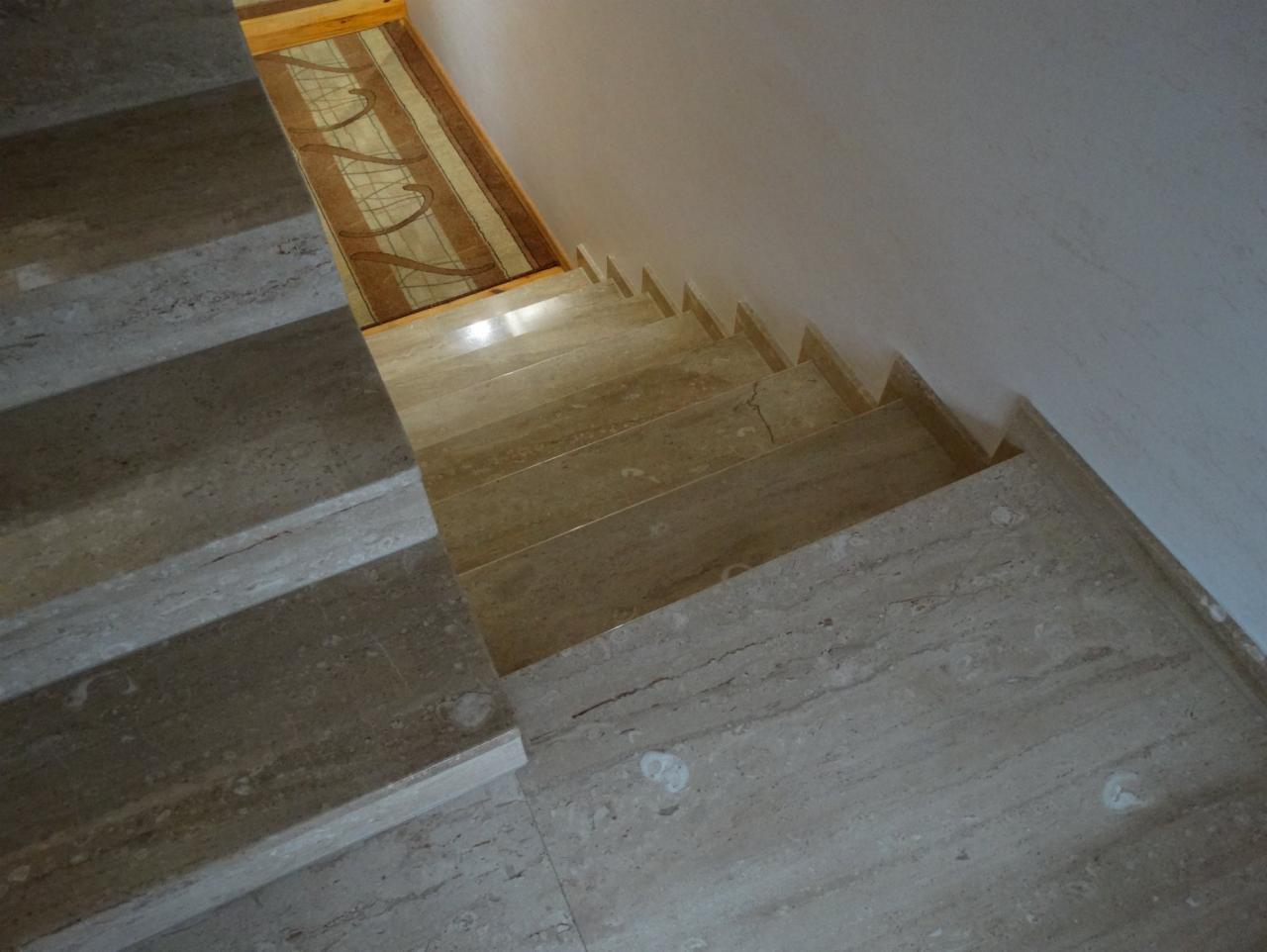 Schody wewnętrzne wyłożone beżowym kamieniem naturalnym z widocznym fragmentem dywanu na podeście, oświetlone naturalnym światłem.