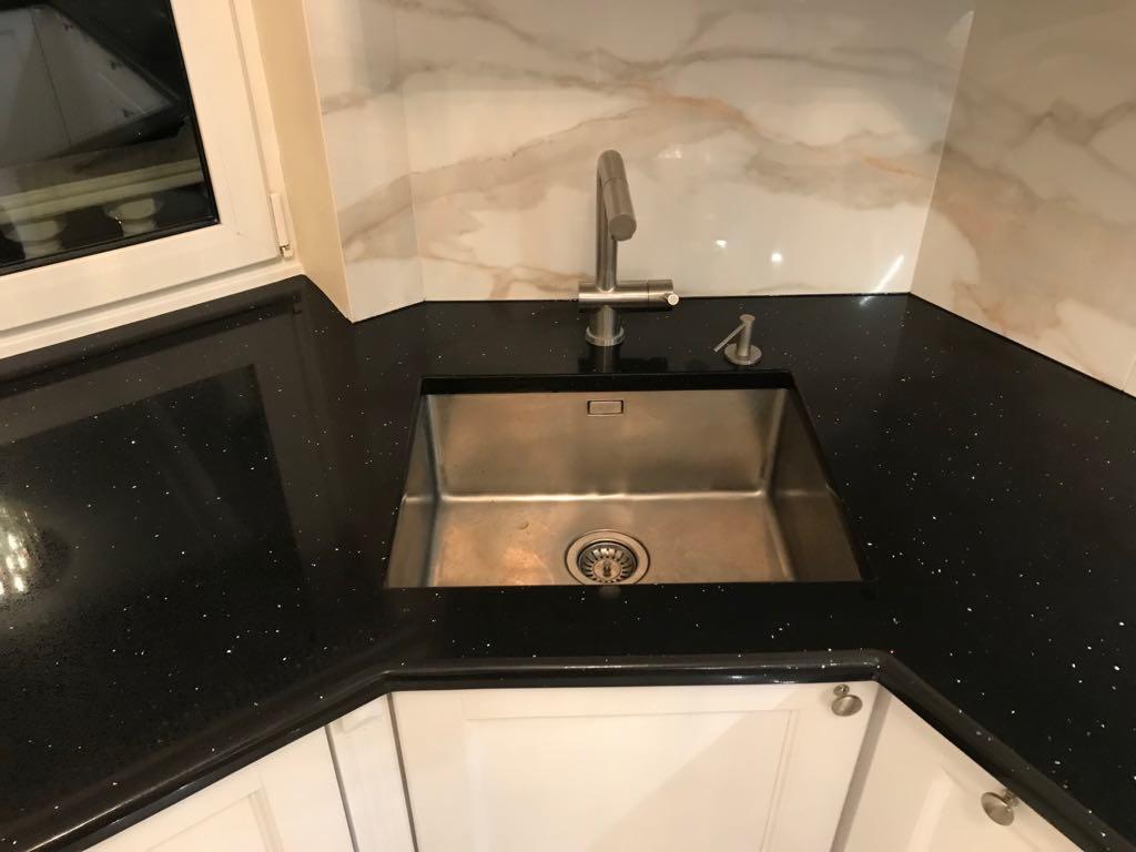 Błyszczący, czarny blat kuchenny z metalowym zlewem i baterią, marmurowe wykończenie ściany w tle, białe szafki na dole.