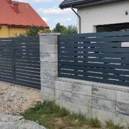 Nowoczesne ogrodzenie posesji z przesuwną bramą, szare metalowe panele poziome na betonowej podmurówce i słupkach z widoczną skrzynką na listy i przewodem elektrycznym.
