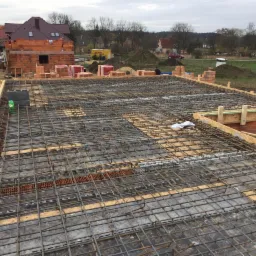 Zbrojenie płyty fundamentowej z widocznymi elementami szalunku i częścią wznoszonej ściany budynku w tle.