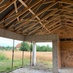 MP Budownictwo Marcin Pokrywka - Wnętrze surowego stanu domu w trakcie budowy: widok na drewnianą konstrukcję dachu z czarną membraną, ściany z czerwonej cegły, betonowe słupy i widok na łąkę z żółtą koparką w tle.